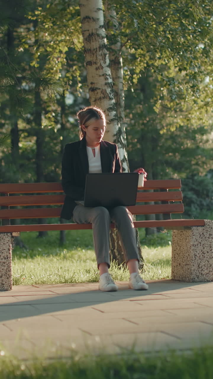 Remote worker seated outdoors on wooden bench using laptop and sipping coffee with lush green trees, sunlight, and passing bus in background