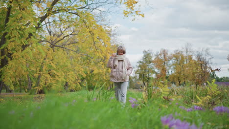 close up of woman walking along autumn garden path as leaves sway gently in wind among purple blooms and golden foliage wearing pink coat, chunky muffler, grey jogger pants and white sneakers