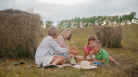 family seated on grass enjoying fresh pastries from picnic basket, young boy in green shirt eats immediately while girl in pink shirt carefully picks her choice