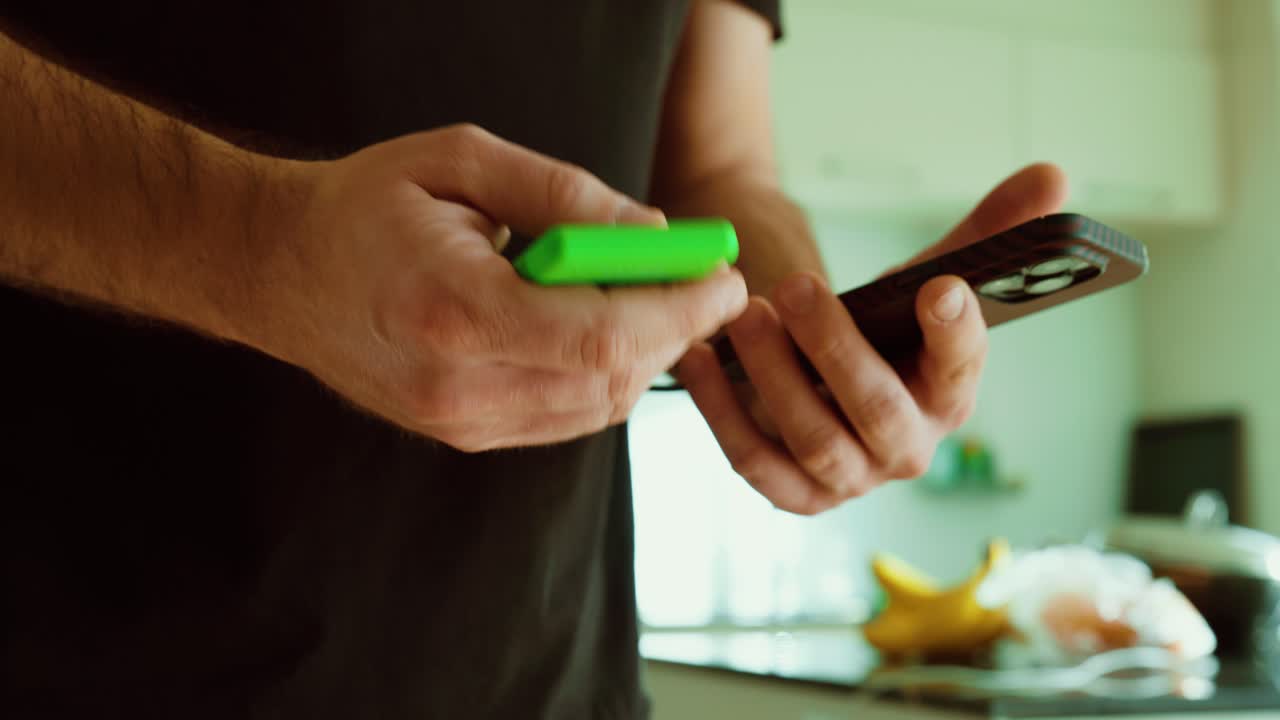 Close-up of a man holding a smartphone and a portable power bank, preparing to recharge the device in a home kitchen setting. Ideal for mobile battery, emergency charging, travel essentials, and every