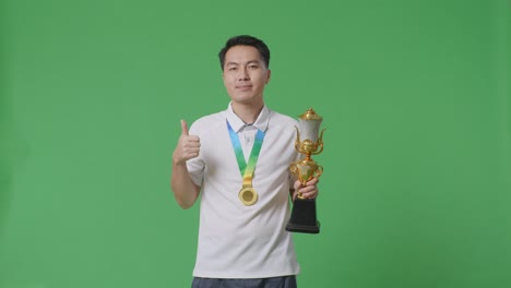 asian man with a gold medal and trophy showing thumbs up gesture and smiling to camera as the first winner on green screen background in the studio