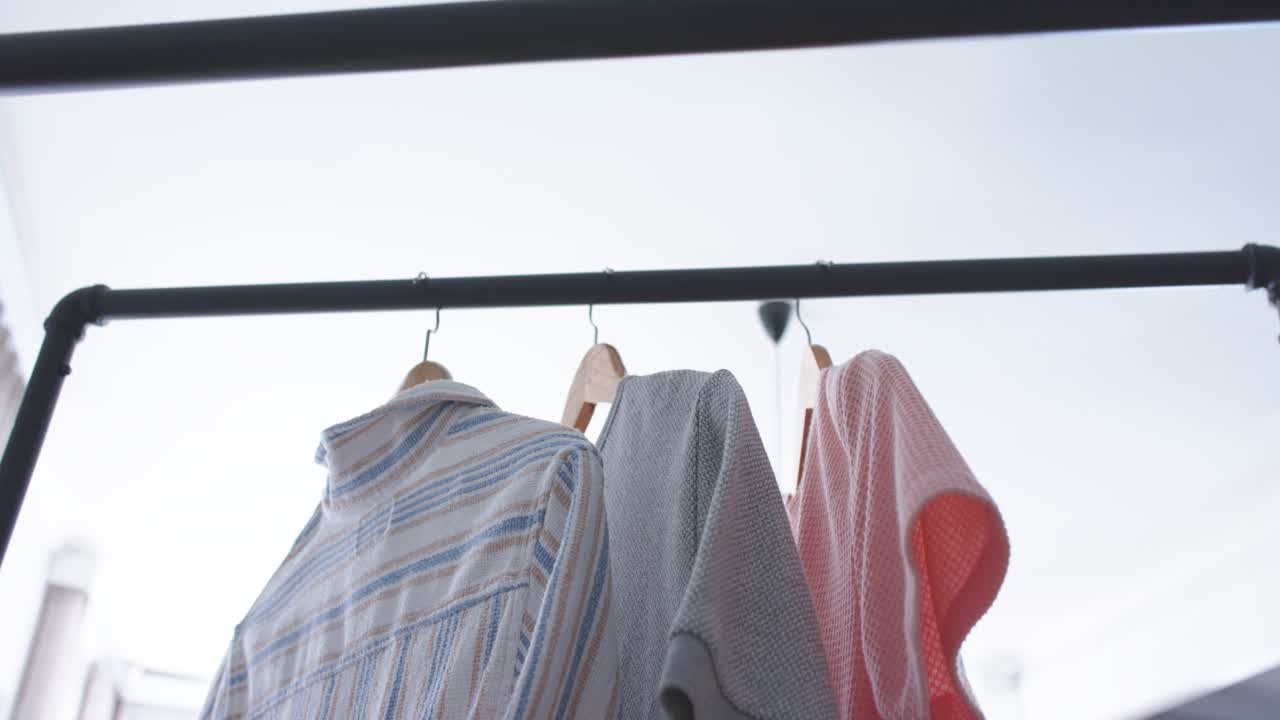 Camicia a righe, maglione grigio e capo in maglia rosa su appendini che dondolano su una barra di metallo nera mentre la telecamera si inclina.