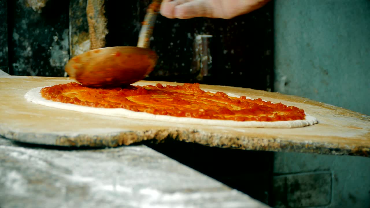 4k, крупный план шеф-повара, распространяющего томатный соус на тесто пиццы с помощью металлической ложки в коммерческом ресторане
