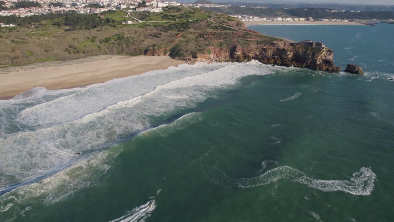 Onde che si infrangono dolcemente sulla riva di una spiaggia deserta a Nazaré e contro un'imponente scogliera che costeggia la costa.