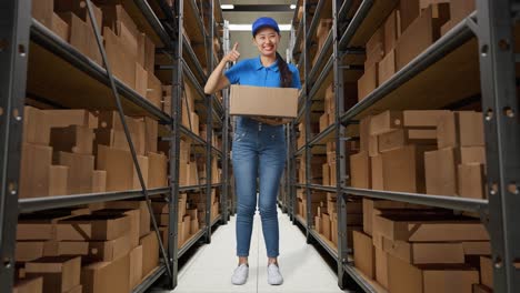 full body of asian female courier in blue uniform showing thumbs up gesture and smiling while delivering a carton in warehouse