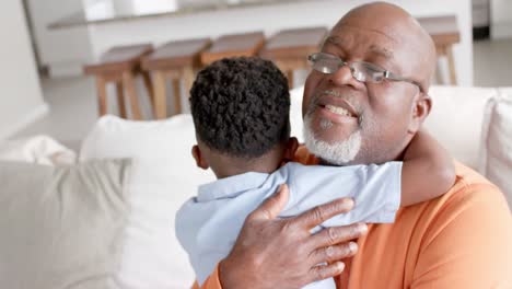 happy african american grandson and grandfather embracing and smiling on couch at home, slow motion