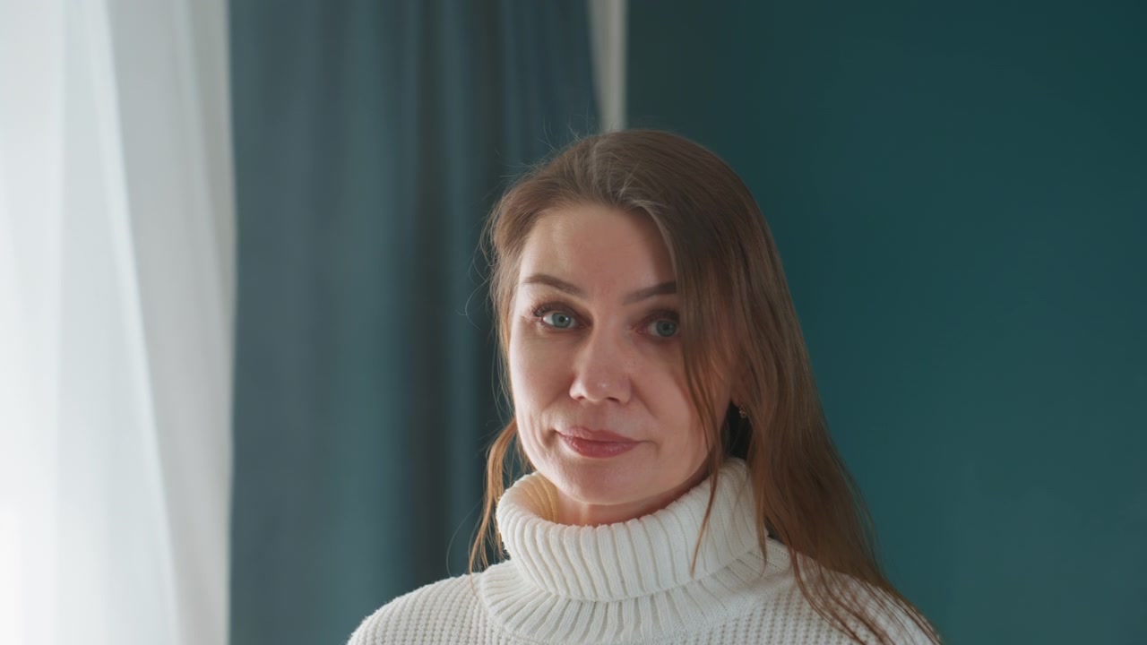 Donna caucasica con maglione bianco che legge appunti di grammatica, sfondo parete color ottanio e tenda trasparente, ambiente accogliente per lezioni private al chiuso con primi piani dell'insegnante concentrata che sfoglia le pagine, sorriso gentile e maglia calda
