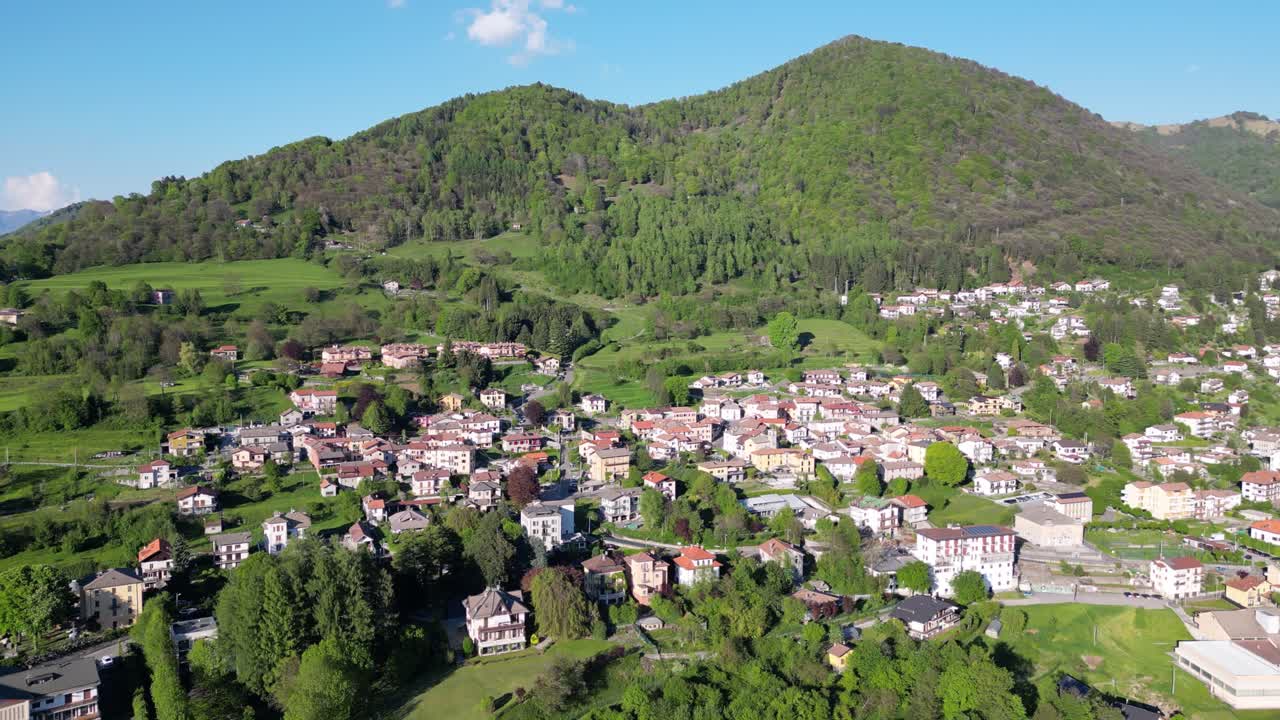 Above San Fedele near Como, traditional rooftops and narrow streets stretch beneath lush green hills and towering mountains, as a drone flies sideways capturing the summer scenery