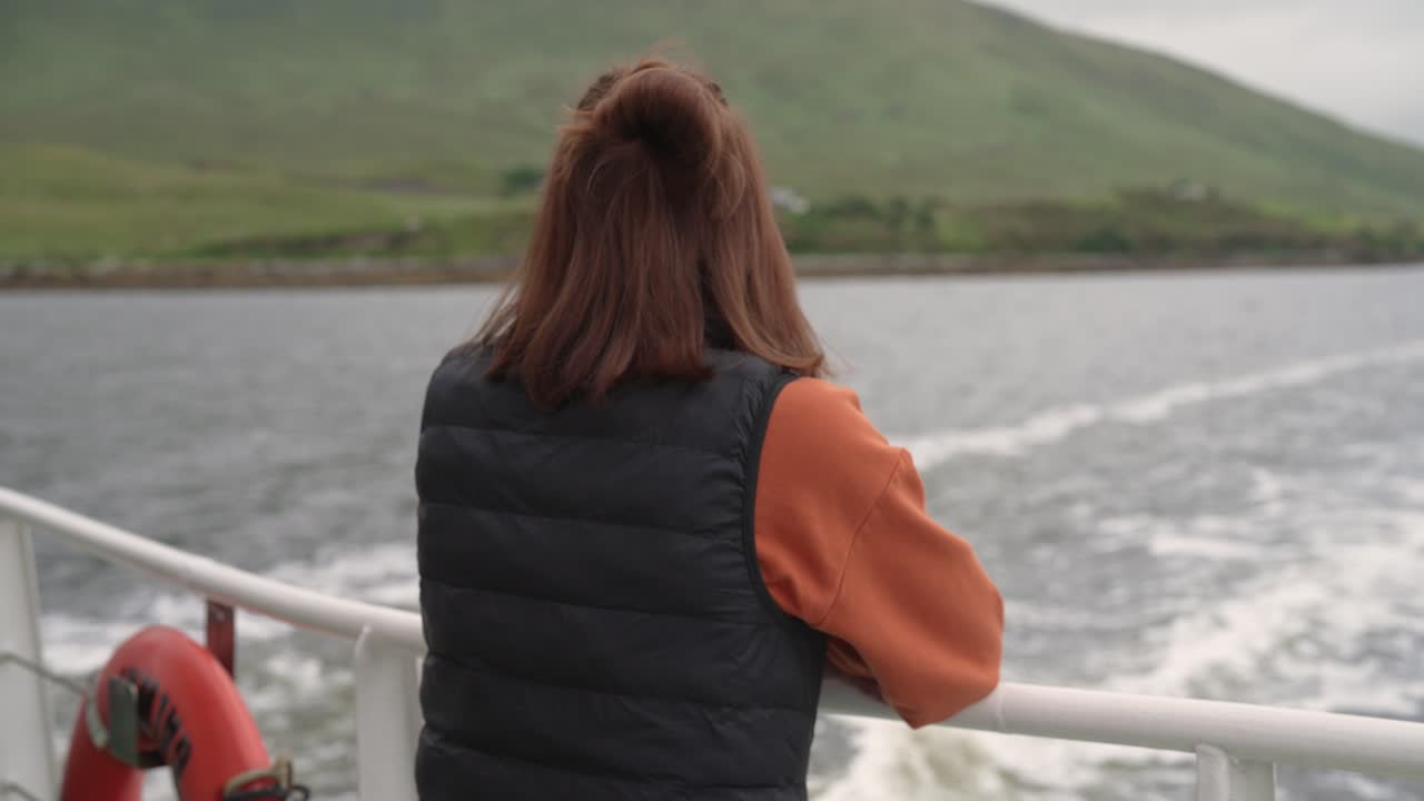 Shot from behind, a woman stands at the back of a boat, watching the wake in the water as it sails away from the Irish coast. A scene of travel, departure, and reflection
