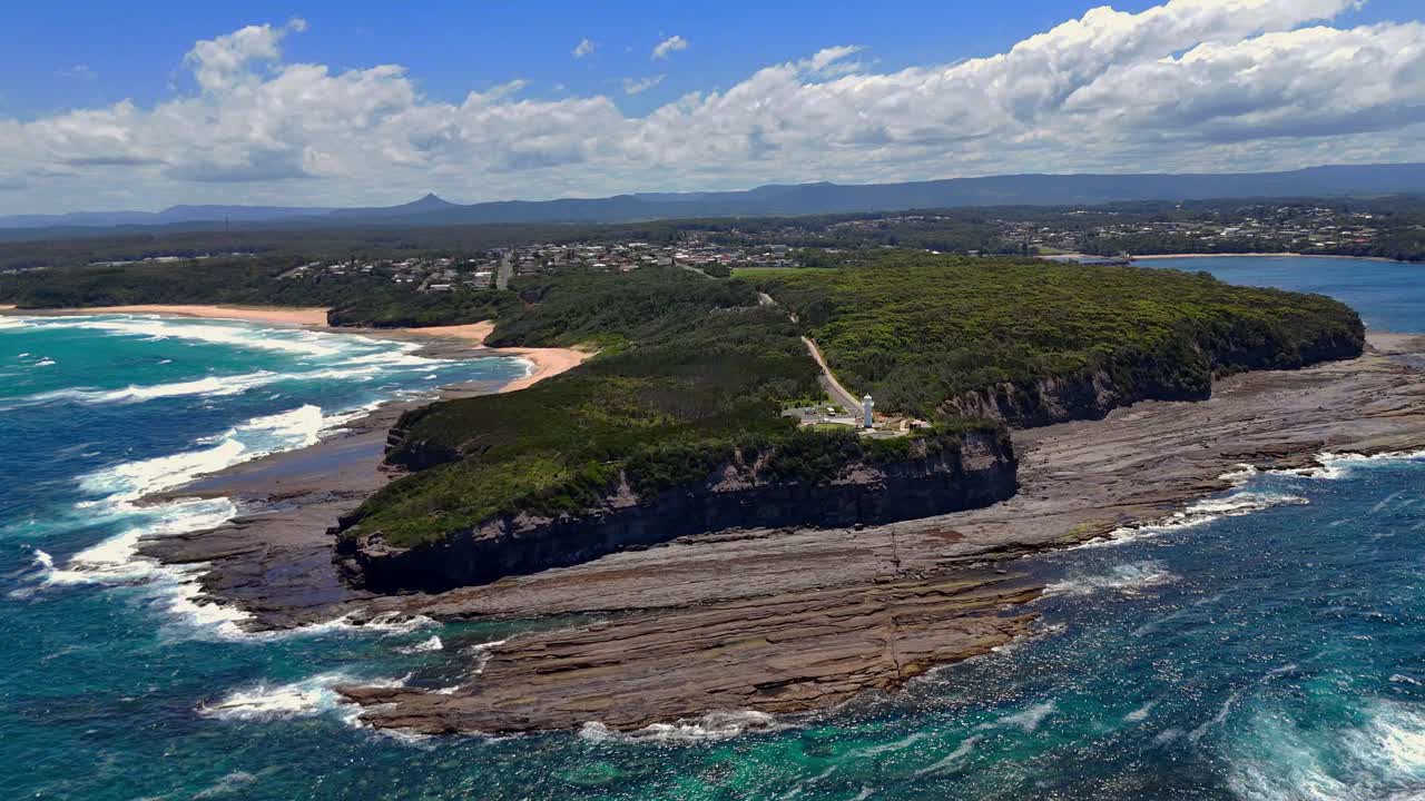 Sweeping establishing aerial of surf waves pounding dark rocks beneath Warden Head Lighthouse on the coastline, vibrant bright sunny day
