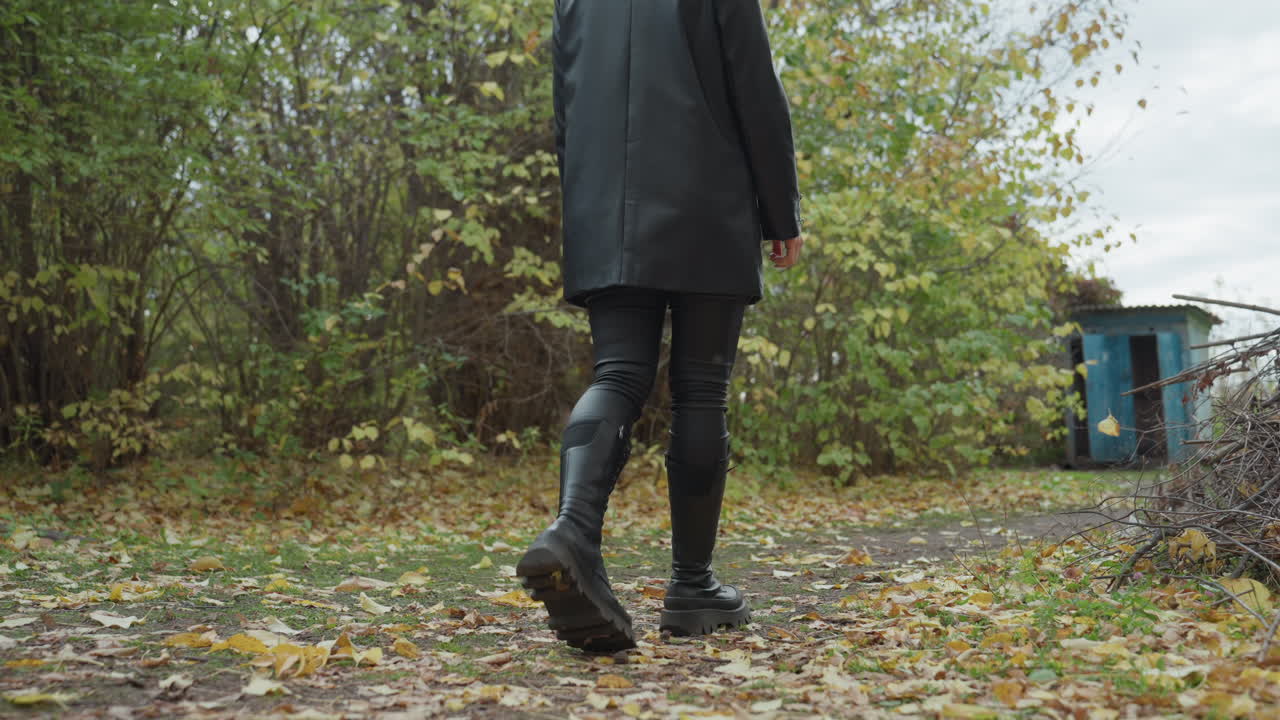 Une femme en veste de cuir se promène dans les bois d’automne sur un chemin jonché de feuilles, portant un pantalon et des bottes en cuir, avec une démarche lente et assurée vers un petit cabanon bleu sous un ciel nuageux, ambiance sombre et granuleuse.
