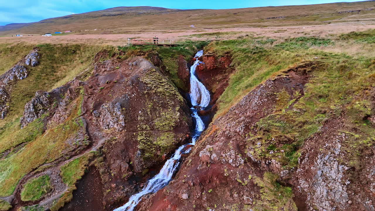 Two people stand on the observation deck located on the mountain. Tourists watch the waterfall flows by the rocks. Top view. Iceland landmarks.