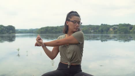 hermosa mujer hispana con anteojos sentada en posición de meditación junto al agua, estirándose y respirando en un entorno tranquilo y pacífico practicando una buena rutina de salud