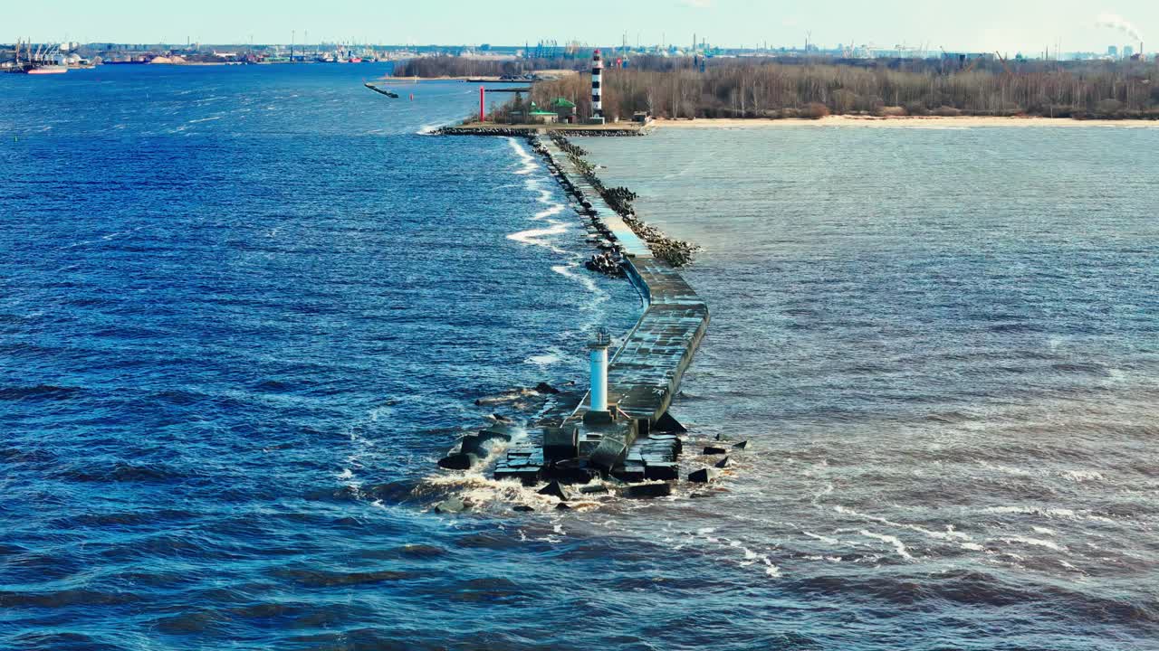 A long stone jetty cuts through vibrant blue sea toward a black and white lighthouse, dividing the choppy water from a calm river estuary and distant forested coastline.