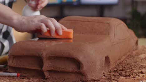 close up of man automotive designer's hands spraying and using sponge to smooth out the surface of the sculpture of car clay in the studio