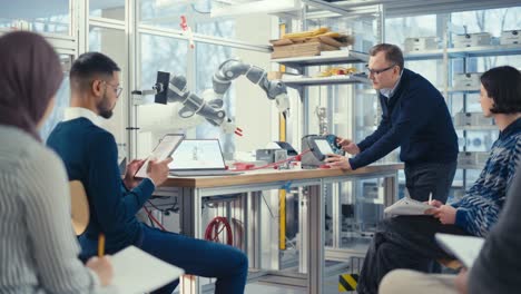 ingeniero jefe masculino manipula con brazo robótico y habla con jóvenes especialistas en computadoras en la sala de reuniones de la fábrica, encuentran soluciones a problemas. desarrollo y concepto de robótica