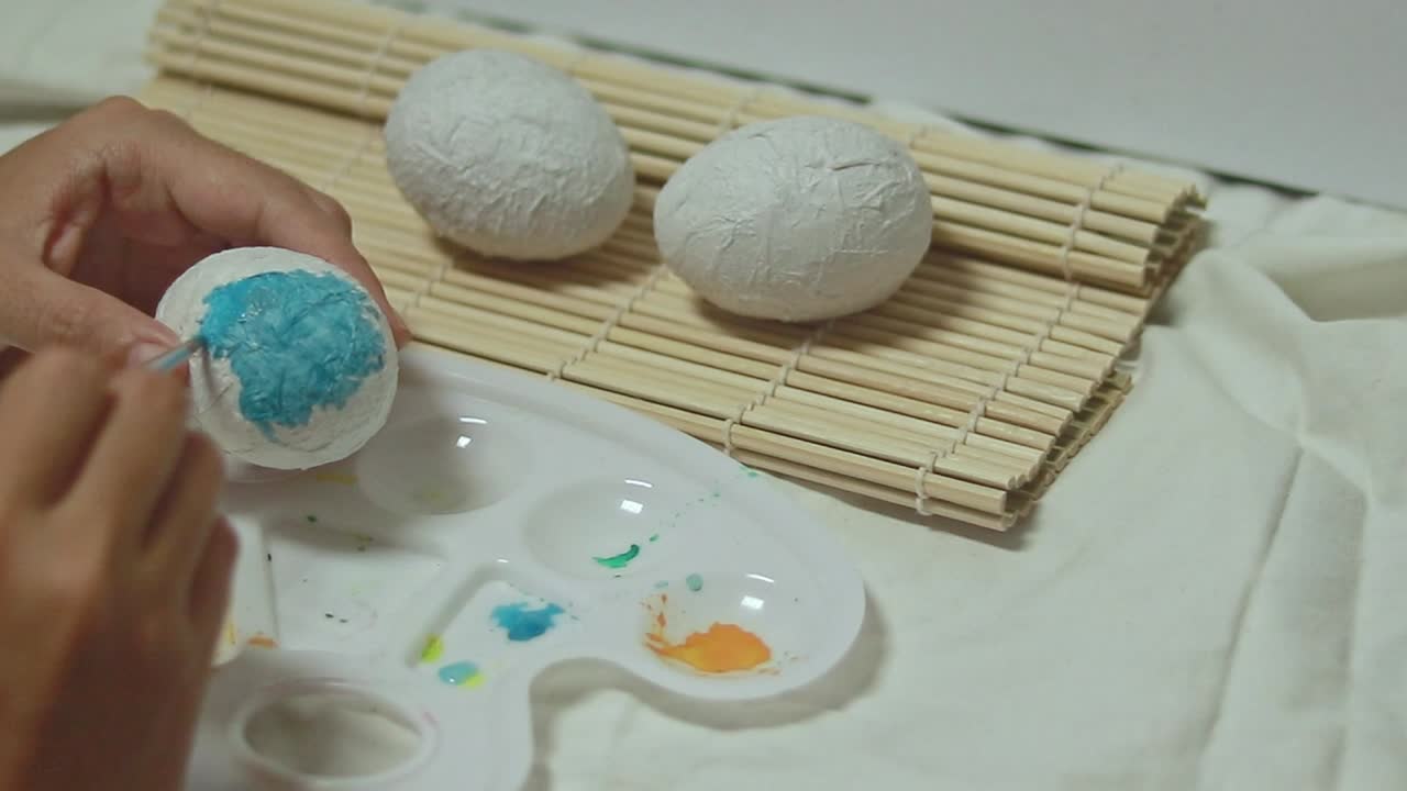 A hand paints a paper mache egg with blue with watercolor, easter egg recycling craft project for Spring season