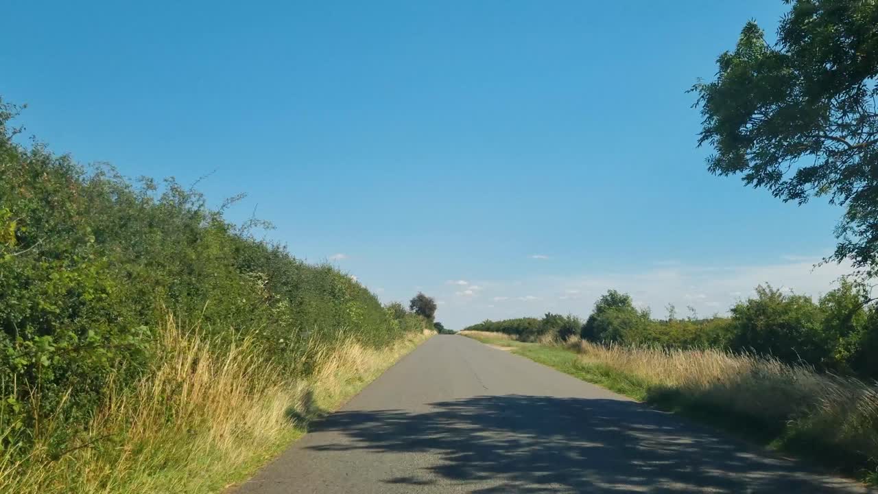 View from a car driving a narrow country road in rural Britain, passing green fields and farmland in a peaceful countryside landscape