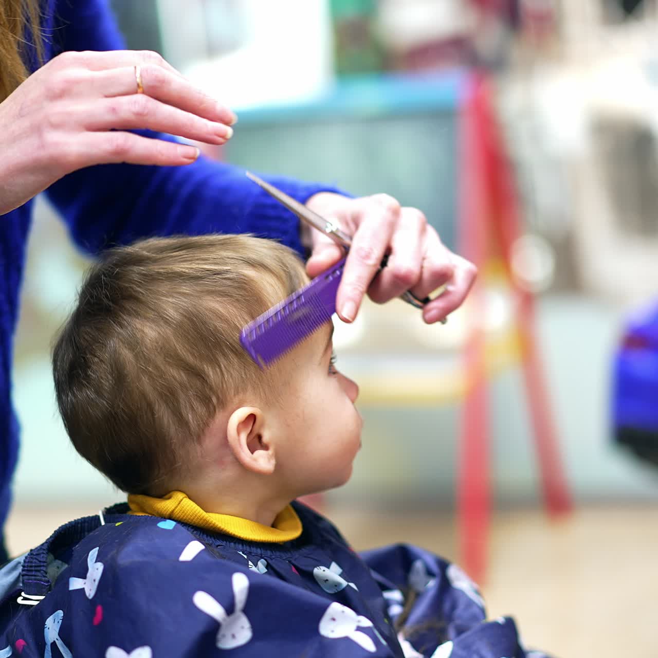 Cutting hair for one year old toddler in a salon. Long-haired barber in blue sweater cuts little boy's hair wearing a cape