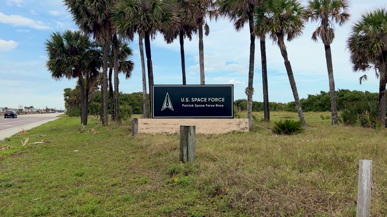 Wide static shot of Patrick Space Force Base main entry sign, centered, on a sunny Florida day. Traffic flows left, palm trees sway, vibrant coastal scene, clear sky, warm summer vibe, military base.