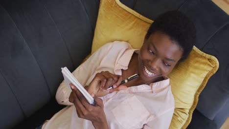Feliz-Mujer-Afroamericana-Tumbada-En-El-Sofá-De-La-Sala-De-Estar,-Escribiendo-En-Un-Cuaderno