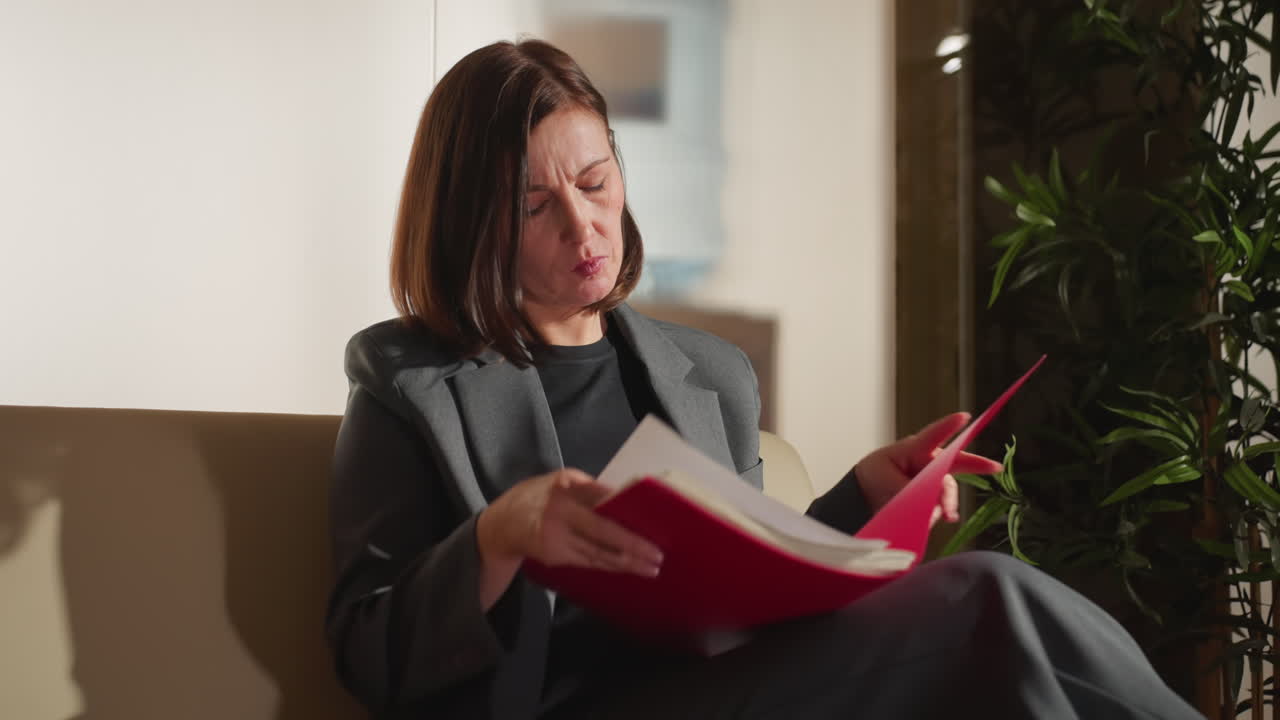 Focused businesswoman in gray suit closely examining papers in red folder while seated indoors, appearing serious and concentrated, surrounded by natural light and office elements