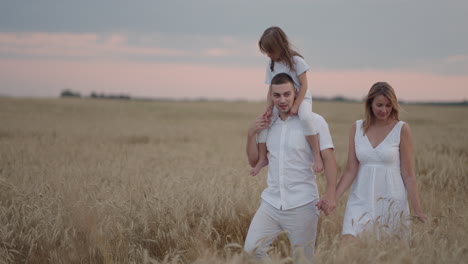 Junges-Elternpaar-Mit-Mädchen,-Die-Sich-An-Den-Händen-Halten-Und-Bei-Sonnenuntergang-Durch-Das-Weizenfeld-Laufen.-Glückliche-Familie-Joggt-Auf-Der-Gerstenwiese-Und-Genießt-Gemeinsam-Die-Natur.-Zeitlupe