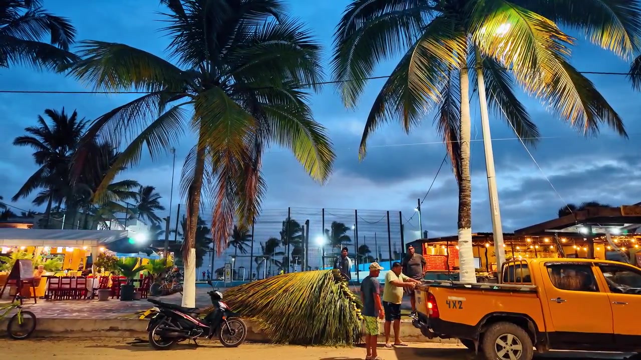 Santo Tomas, Ecuador, 1 maggio 2026: Scena notturna del villaggio delle Galapagos con palme, camion arancione al crepuscolo. Scena di strada all'ora blu, fasci di paglia di palma, ristorante illuminato, palme da cocco, Isabela Galapagos