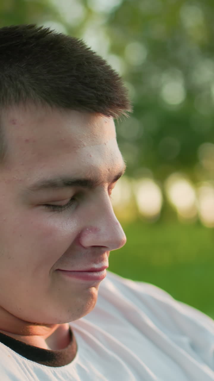 Close up of music artist wearing headphones, nodding head and enjoying music with a subtle smile while looking up, background features vibrant greenery, trees, and sunlight