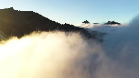 Volando-Por-Encima-De-Las-Nubes-Bajo-Los-Picos-De-Las-Altas-Montañas-Bajo-El-Cielo-Despejado-De-Verano-Y-La-Luz-Del-Sol-De-La-Hora-Dorada,-Disparo-De-Drones