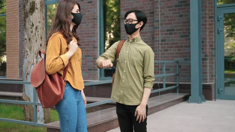 보호용 얼굴 마스크를 쓰고 대학 근처 거리에서 팔꿈치로 인사하는 아시아 남자 학생과 백인 여자 학생