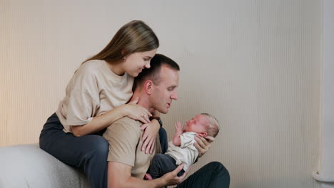 lovely young caucasian family of three. man hold a baby sitting on sofa and woman sits above him embracing husband.