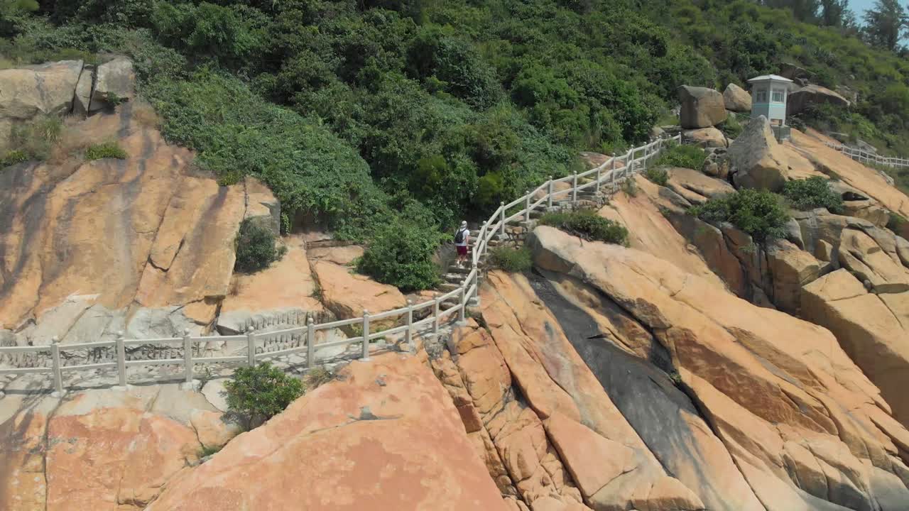 바위가 많은 해안 트레일을 따라 배낭을 메고 걷는 남자의 공중 공개 샷을 축소합니다.