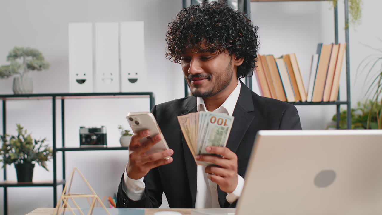 Happy rich indian businessman counting money dollar cash use smartphone calculator app at office