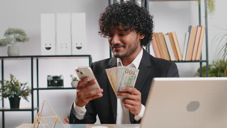 happy rich indian businessman counting money dollar cash use smartphone calculator app at office