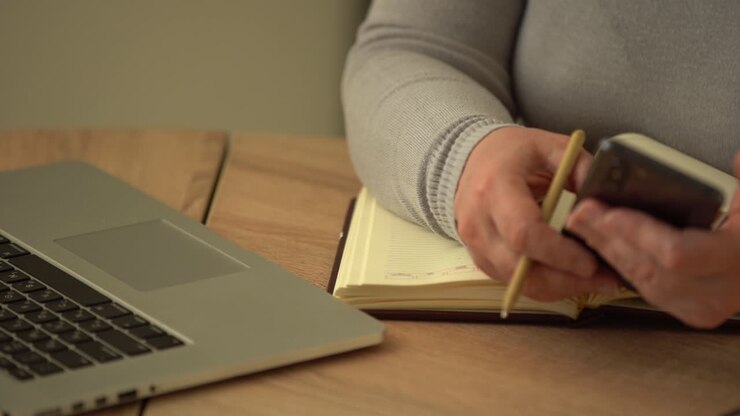 Close up wrinkled male hands writing information. Old mature woman working at office, using computer, handwriting notes
