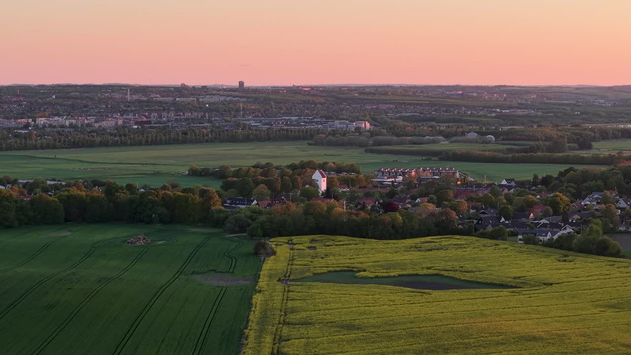 Aerial drone view of Danish countryside at sunset with green fields, yellow rapeseed crops, and a distant city skyline