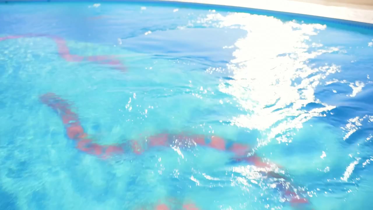 Permukaan kolam renang yang tersinari matahari menunjukkan gerakan air yang lembut dengan sorotan berkilauan dan garis merah terdistorsi bergerak di bawah air biru jernih, ditangkap dari atas dengan pola reflektif cerah.