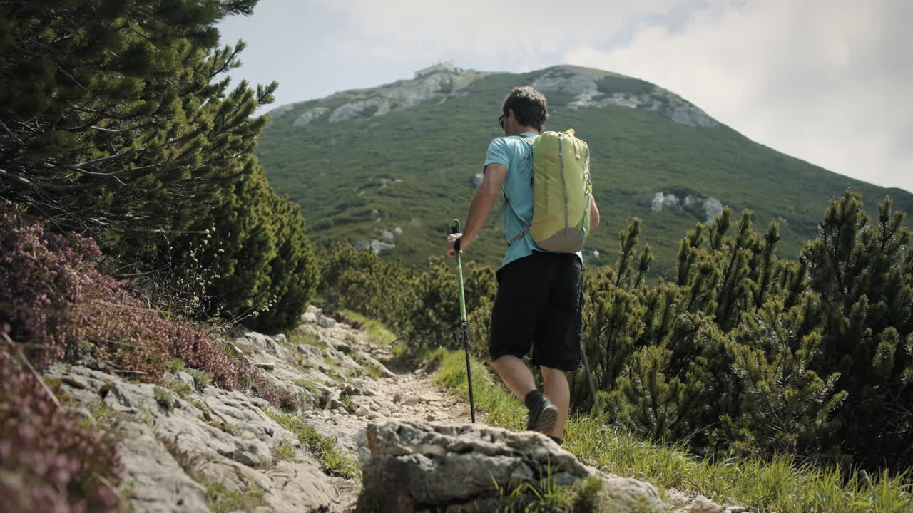 seguimiento de cámara detrás de un excursionista con bastones de senderismo y mochila verde claro que camina por un camino rocoso hacia la cima de la montaña snežnik