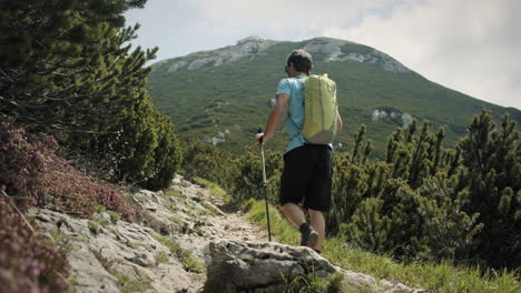 camera tracking from behind a hiker with a hiking poles and light green backpack who is walking up on a rocky path towards the top of the mountain snežnik