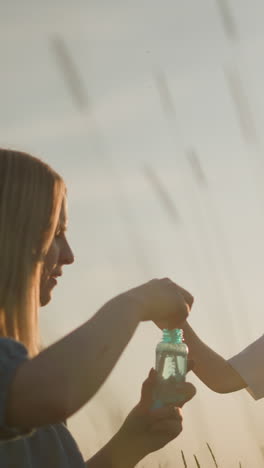 a woman in a blue dress kneels in a grassy field at sunset, holding a bubble bottle as she and her young son blow bubbles from wands. highlighting the bond between mother and child