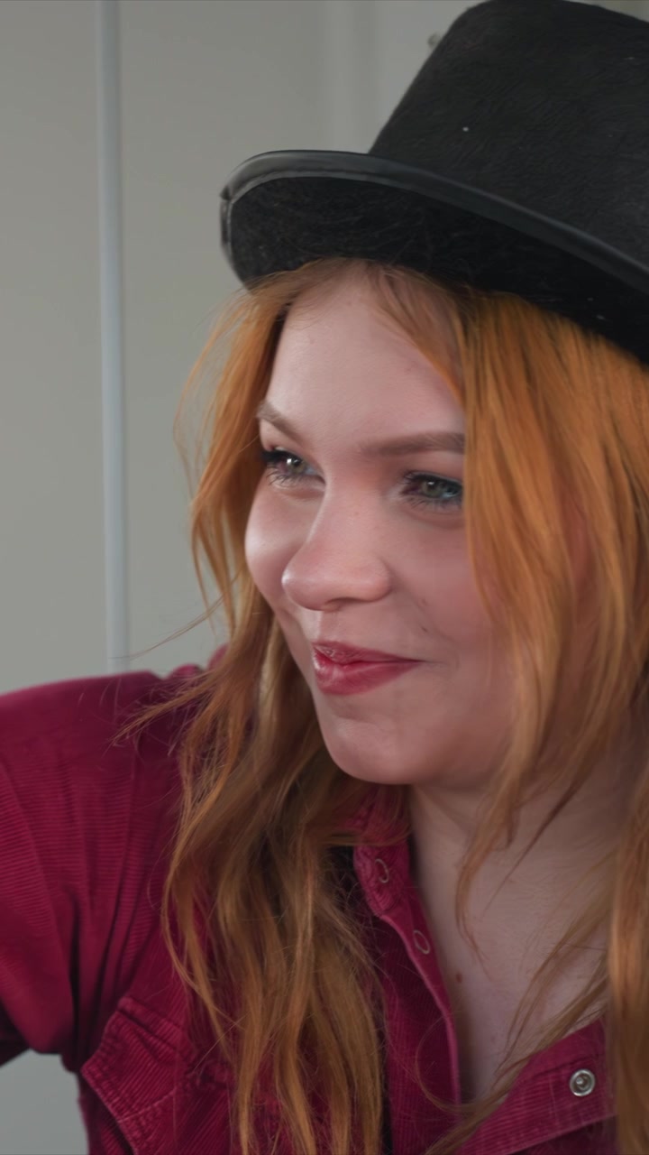 Close up of young woman with long wavy red hair wearing. Her clothing details include visible buttons. Background is plain light colored indoors suggesting indoor