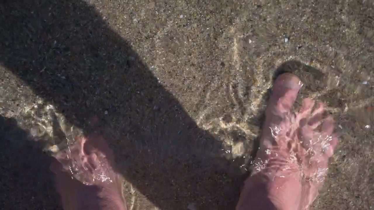 Inquadratura in soggettiva di piedi che stanno in acqua di mare bassa mentre onde leggere si infrangono sulla spiaggia sabbiosa