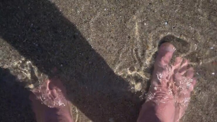 Inquadratura in soggettiva di piedi che stanno in acqua di mare bassa mentre onde leggere si infrangono sulla spiaggia sabbiosa