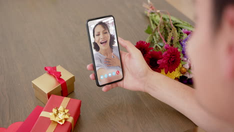 mujer caucásica sosteniendo un teléfono inteligente con mujer caucácea en la pantalla, regalos en el escritorio