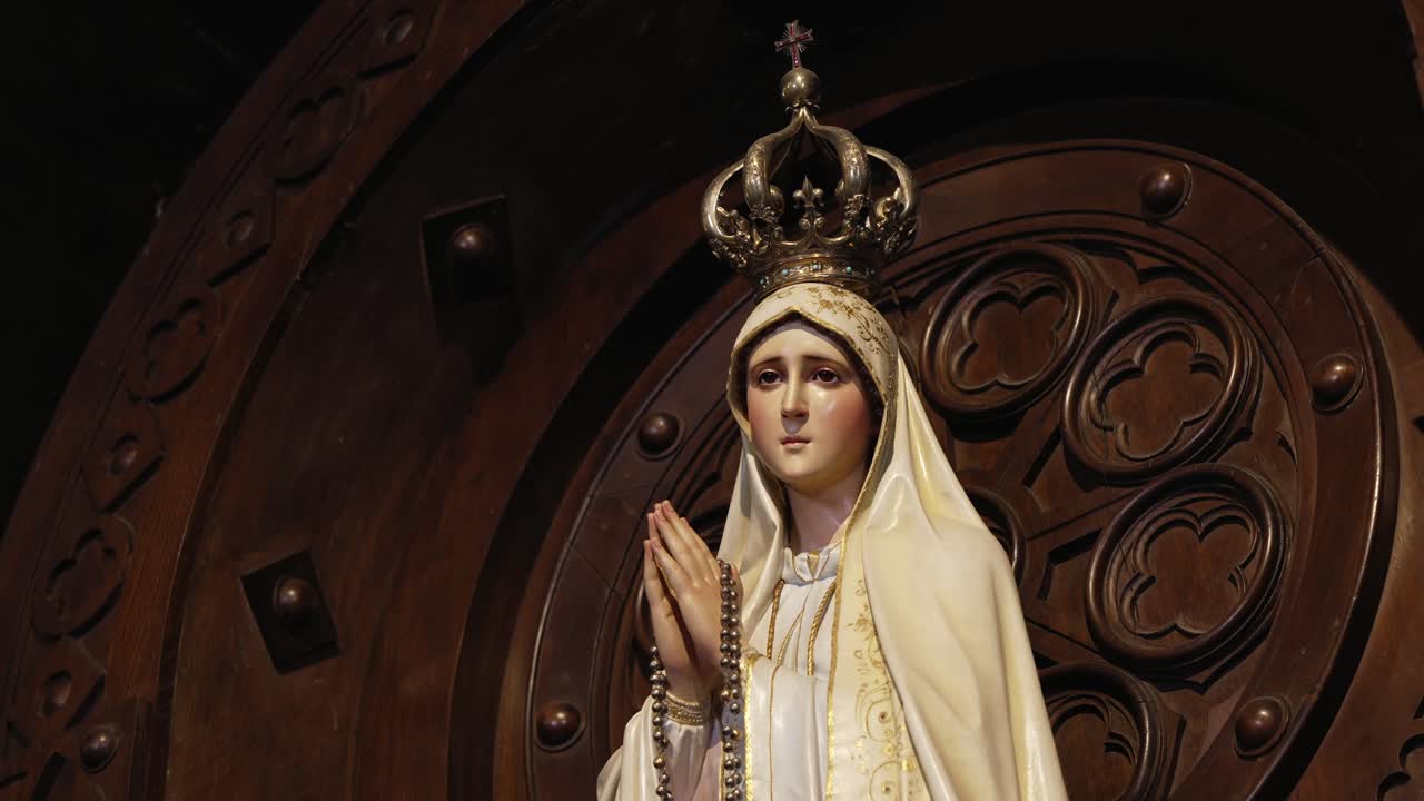 Statue of Virgin Mary with a crown, holding a rosary and in a prayerful pose, against a carved wooden backdrop