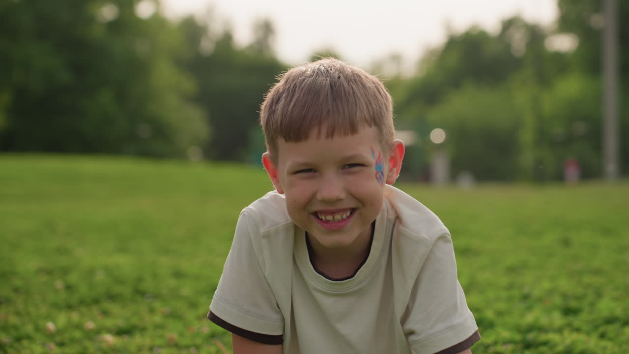 toddler boy smiling while seated on grass, summer park backdrop with soft light, joyful expression capturing carefree outdoor moment, closeup portrait of healthy happy child enjoying nature