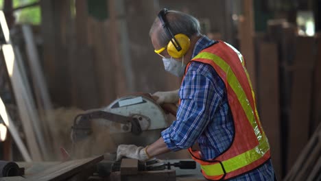 old asian carpenter using circular saw cutting wooden plank in workshop . young man builder sawing board . senior craftsman wearing protective mask , hearing protectors and safety glasses .