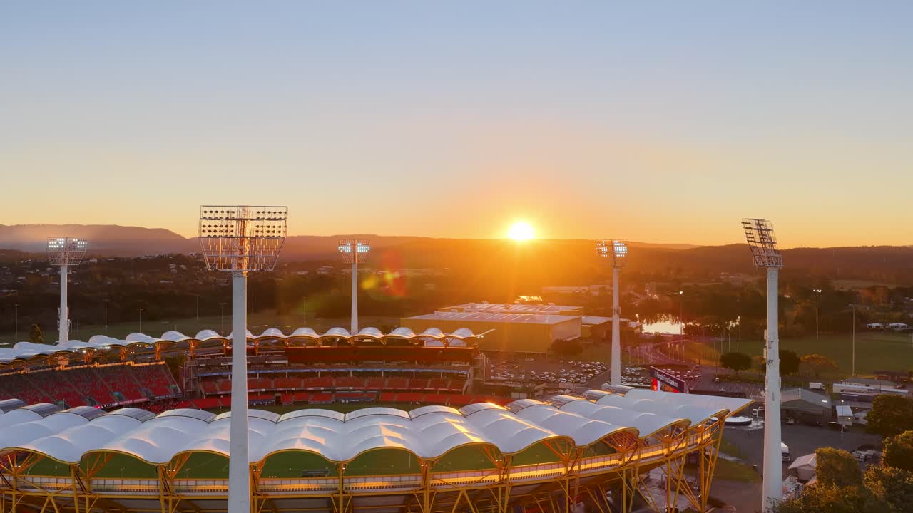 Drone footage glides over a large outdoor stadium at sunset, capturing illuminated floodlights, vibrant seating, and the surrounding landscape under golden hour lighting