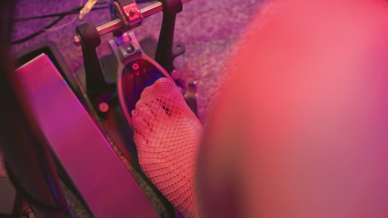 Close up of person leg wearing fishnet pantyhose pressing down drum kick pedal barefoot during studio practice, surrounded by cables and soft carpet under warm colorful lighting
